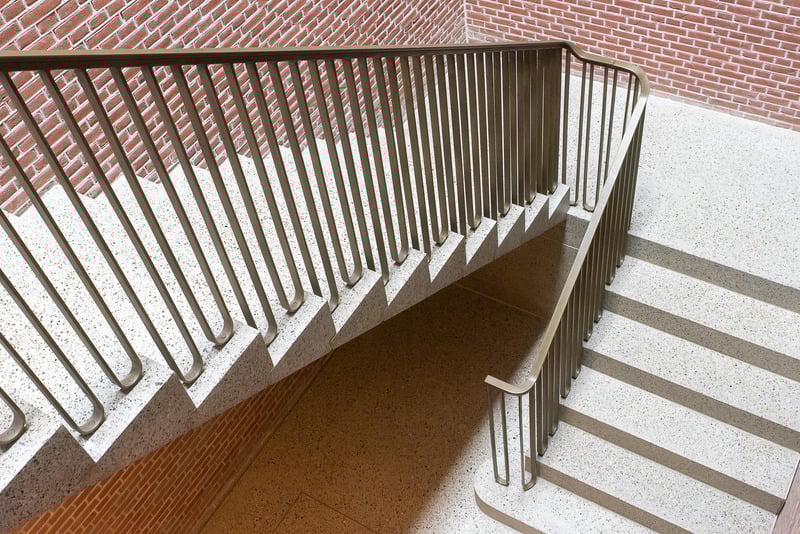 An interior view of a modern stairwell with speckled concrete stairs and landings, brick walls, and brown metal railings.